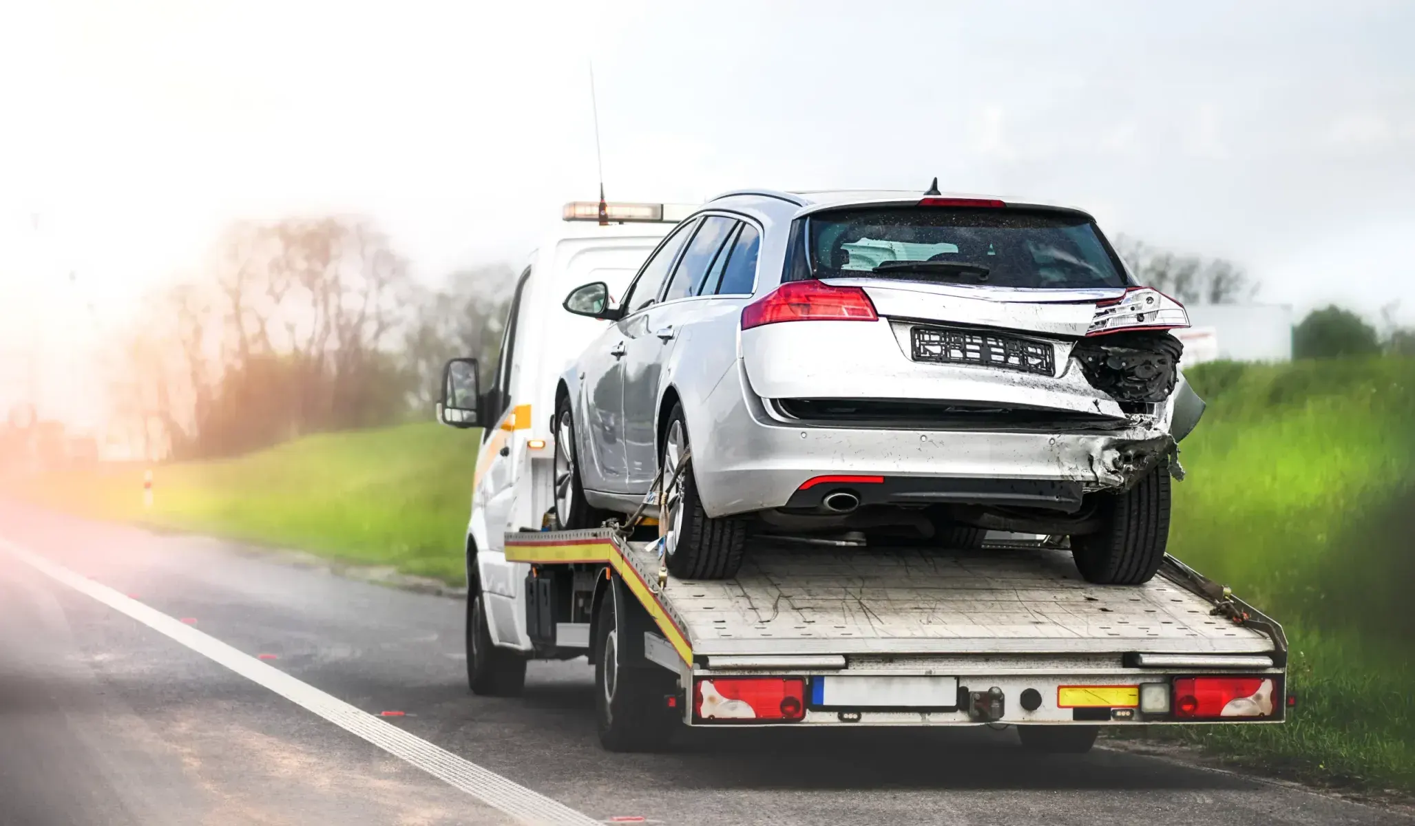 Damaged white car on a tow truck beside a roadside after a crash